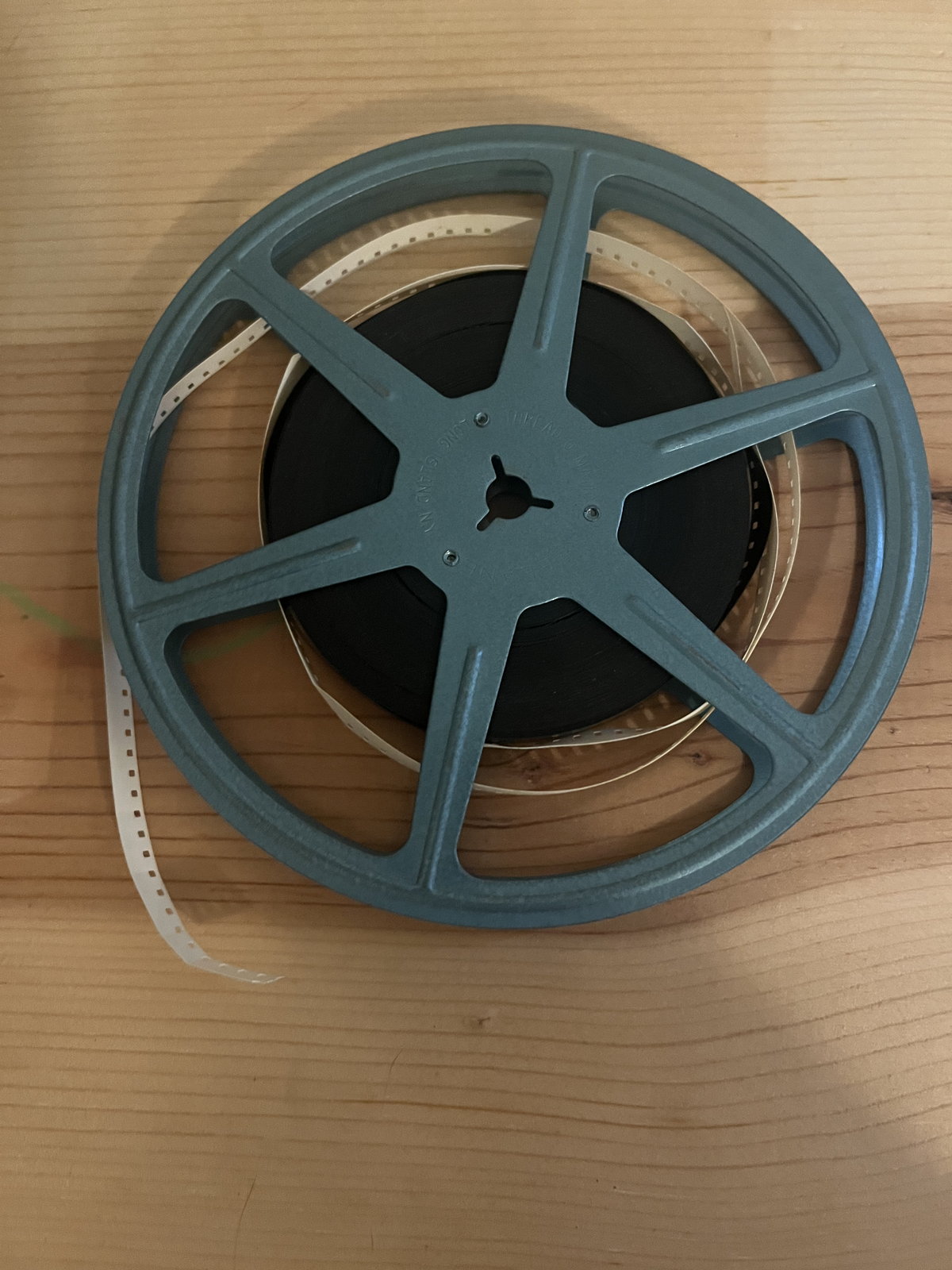 A blue plastic 8mm reel with a short tail of film, lying on a plywood table against a white wall.