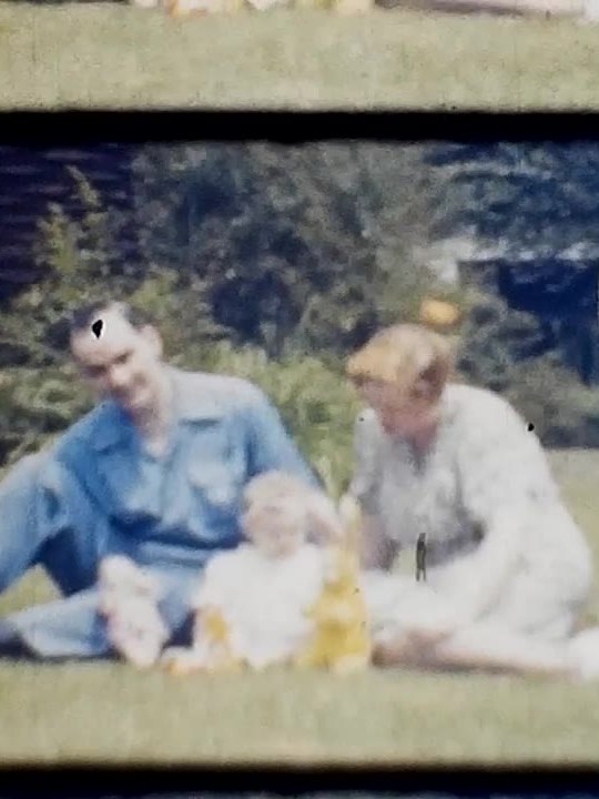 A frame of digitized colour home-movie film: two parents and a baby sitting on a lawn, the film’s frame edges visible.