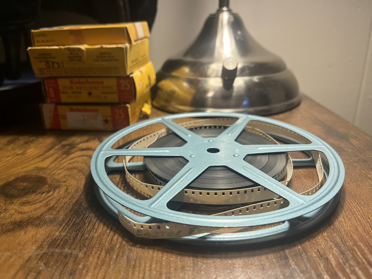 A blue reel of 8mm film lying on a desk under a brass lamp, stacks of film processing boxes behind it.
