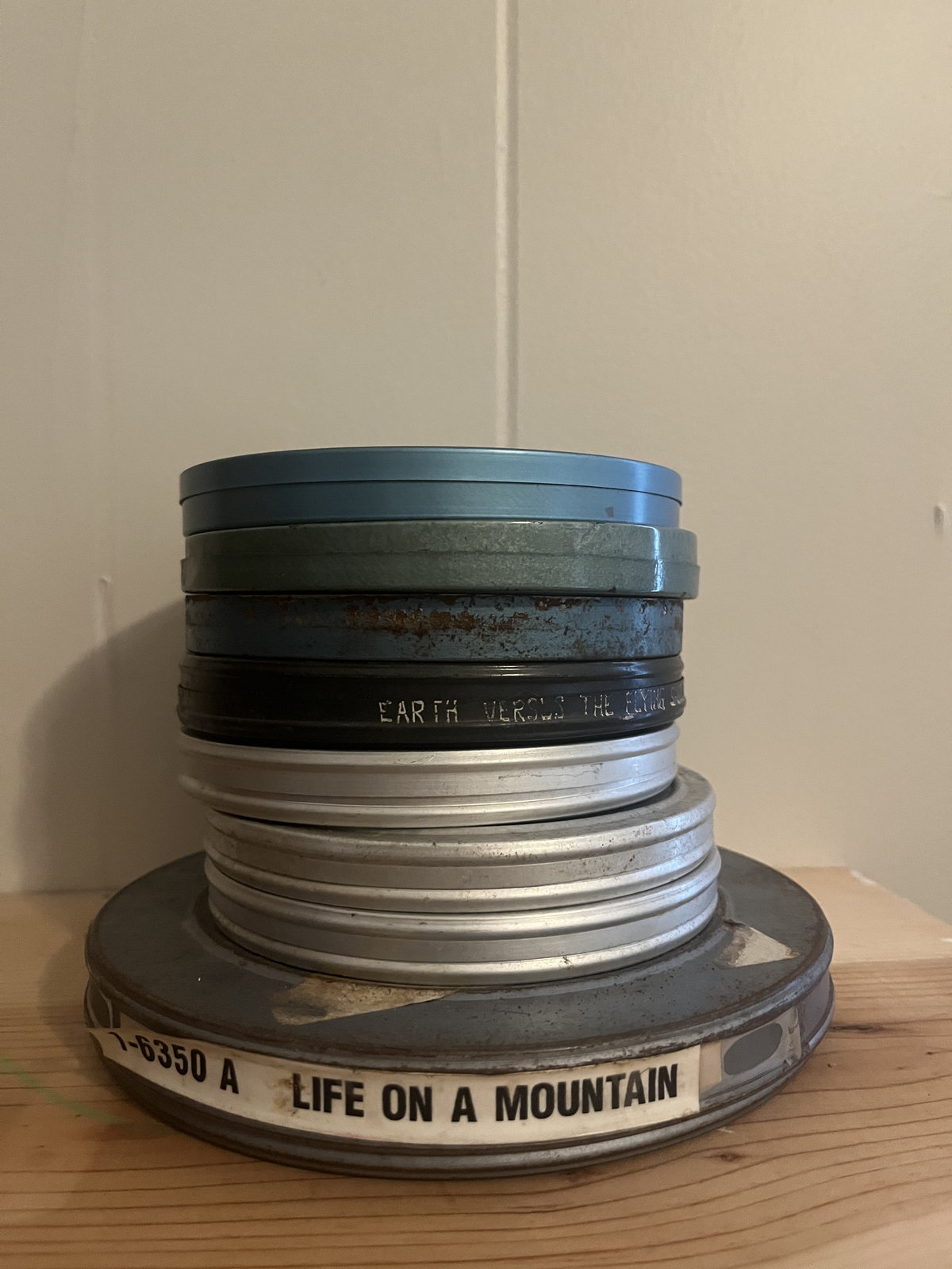 A stack of metal 16mm film cans on a plywood shelf; the bottom can's label reads LIFE ON A MOUNTAIN.